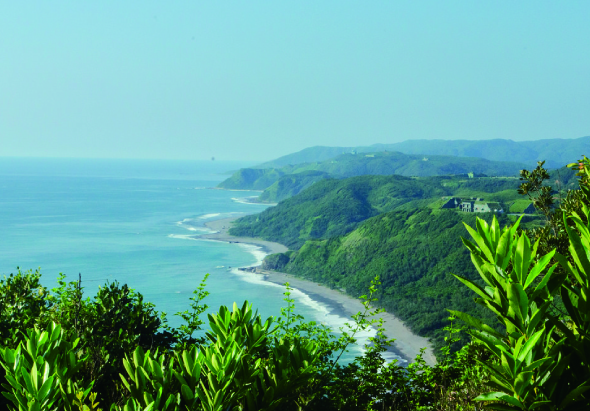 翠綠山丘俯瞰蔚藍海岸線，白色浪花拍打黑沙灘，遠處山脈層疊，晴朗天空映襯熱帶植被，展現自然秀麗的海濱景觀。