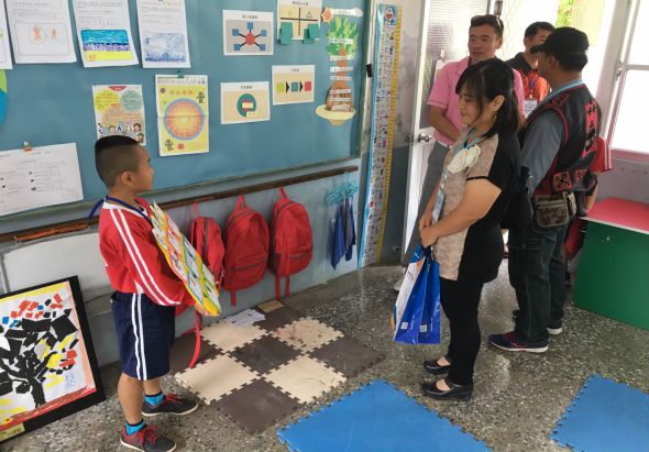 幼兒園教室內，一名穿著紅白運動服的小男孩展示美勞作品，身後牆面貼滿兒童繪畫和教學掛圖。另外五名學童和成人聚集在教室另一側，地面鋪有彩色軟墊，窗邊擺放紅色布偶，呈現教學互動場景。