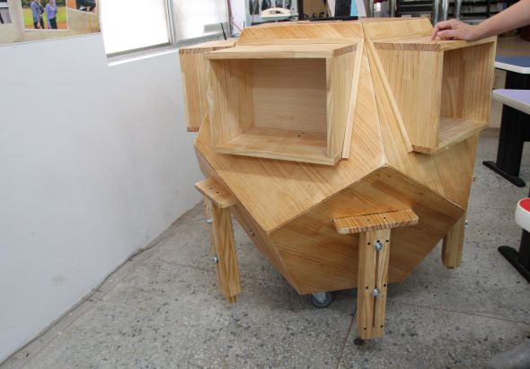 木工工作室內，一張未完成的木製家具作品。淺色木材製作的構件堆疊在帶輪子的木製工作台上，包括空心的木箱結構和平面板材。窗戶和工作環境可見於背景，右側有人員在操作。
