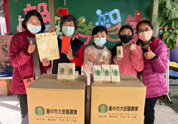 五位戴口罩的人員在綠色佈景前，展示臺中市大安區農會的農產品禮盒。前方擺放兩個印有農會標誌的紙箱，人員舉起拇指和農產品包裝，展現推廣在地農產的活動成果。