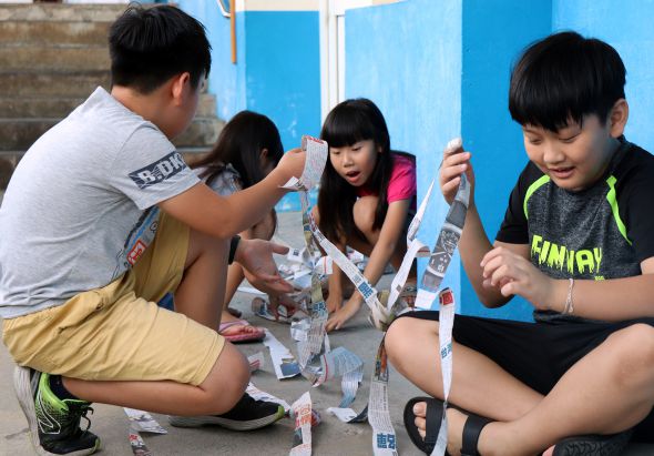 四位年輕人蹲坐在地上，圍成一圈，共同堆疊建築物模型。藍色背景牆後方，他們專注地用白色紙張和材料進行手工創意建築活動，展現團隊合作和創意學習的氛圍。