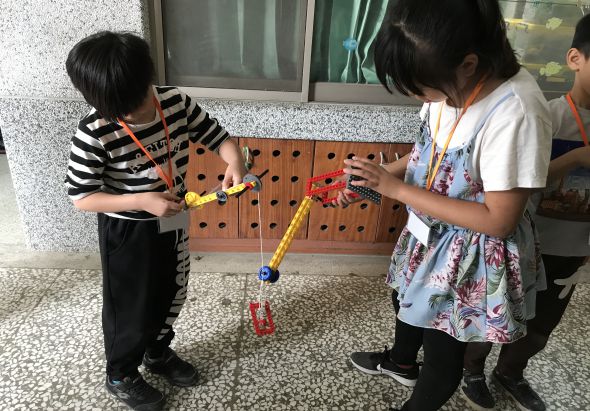 兩名小學生在教室內進行科學實驗活動，他們在木製有孔板前展示和組裝帶有紅黃色零件的建構教具，進行動手學習。背景可見窗戶和教室環境。