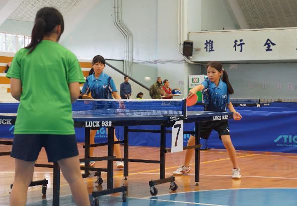 室內乒乓球館中，穿著綠色和藍色運動服的選手們在進行比賽。前景左側選手穿綠衣，對面選手穿藍衣準備迎球。背景可見其他比賽台和觀眾。牆上有中文字樣招牌。