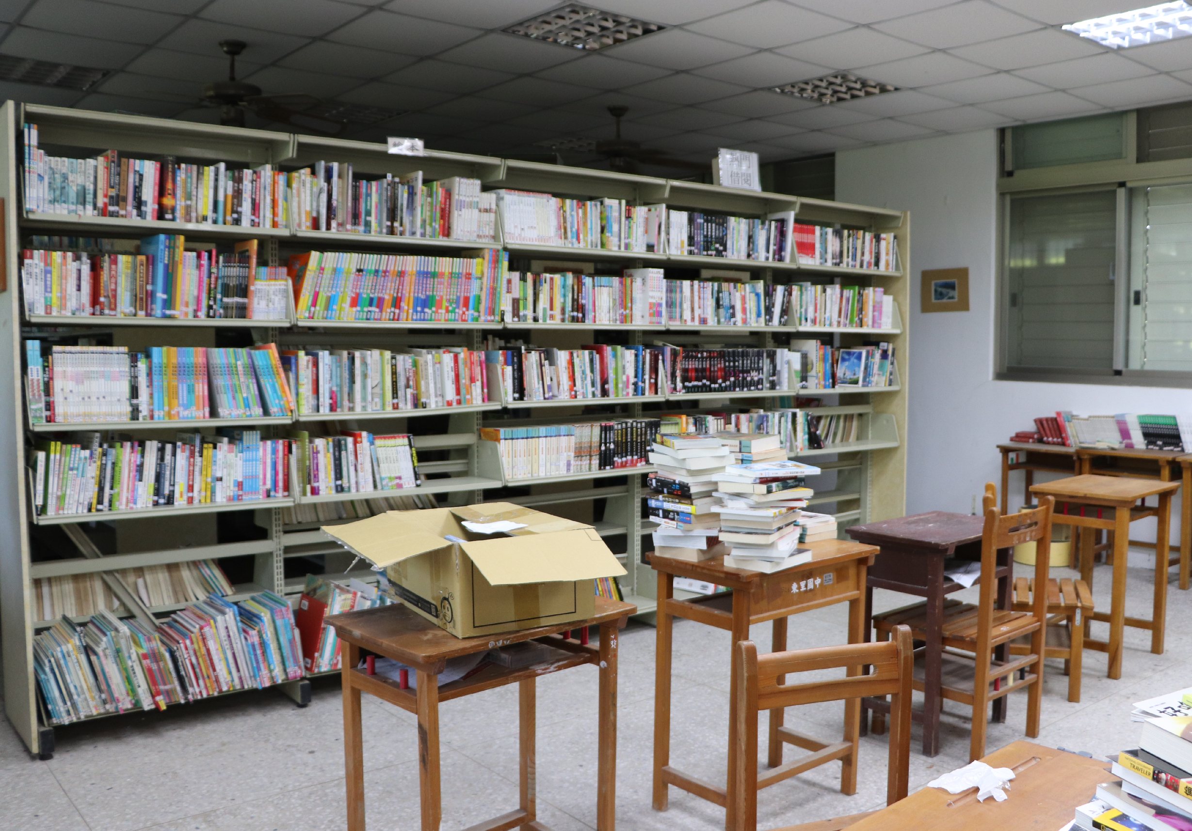 圖書館內部空間，牆面上裝有多層綠色書架，整齊放置各類書籍與雜誌。地面擺放木製閱讀桌椅，桌上堆放待整理的書籍和紙箱。右側角落有小型書架陳列紅色精裝書。天花板裝有照明燈具和風扇，營造靜謐的閱讀環境。