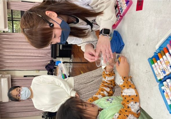 室內教室裡，一名戴著藍色口罩的女性教師正向一名穿著橙色老虎紋衣服的小女孩示範科學實驗，使用透明玻璃杯進行操作。背景有另外兩名女性觀看，桌上擺放著彩色筆盒和實驗用具。
