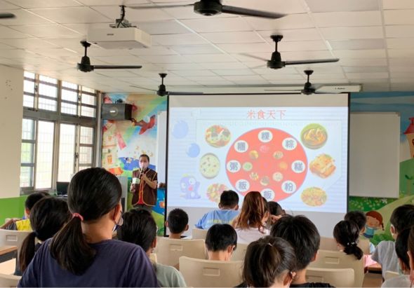 教室內一群學生面對投影螢幕聽課。螢幕上顯示「小食天下」主題，展示多個食物圖片排列成圓形。教室配備天花板吊扇、窗戶和彩色教學布置。