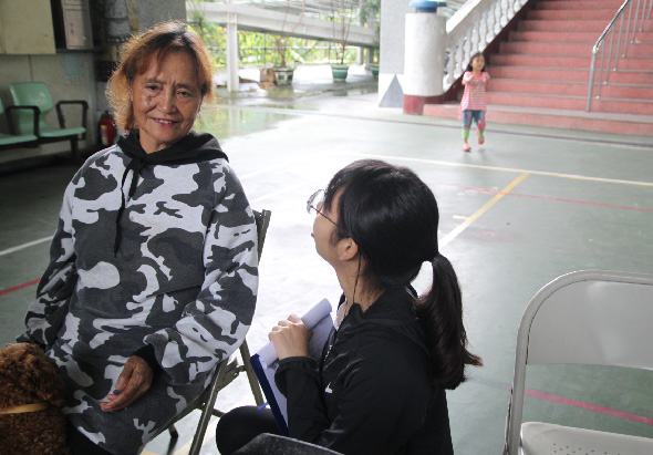室內籃球場，一位穿著黑白迷彩夾克的年長女性站立，一位穿著黑色服裝的年輕女性坐在椅子上面向她，兩人進行交談。背景可見紅色樓梯和其他學生活動。