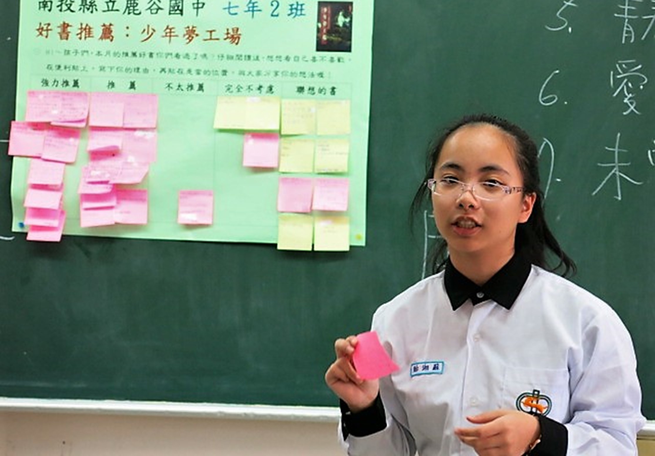 國中女學生在教室前展示專題海報，海報標題為「好書推薦：少年夢工場」，貼有多張粉紅色和綠色便利貼。學生穿著校服，手持粉紅色卡片，背後黑板上有中文字跡。