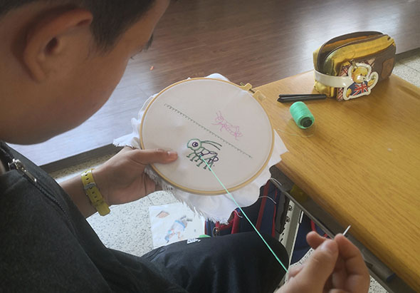 一名學生在木製桌上進行刺繡練習，手持繃圈上的布料，正用針線進行刺繡作業。桌上擺放著黃色膠帶、綠色筆和其他手工藝材料，展示傳統手工藝學習的情景。