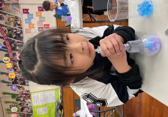 小女孩在教室內進行科學實驗，她專注地觀察裝有藍紫色液體的透明瓶子。桌上放有玻璃杯和實驗用具。背景可見教室布置、學生作品和彩色字母牆飾。