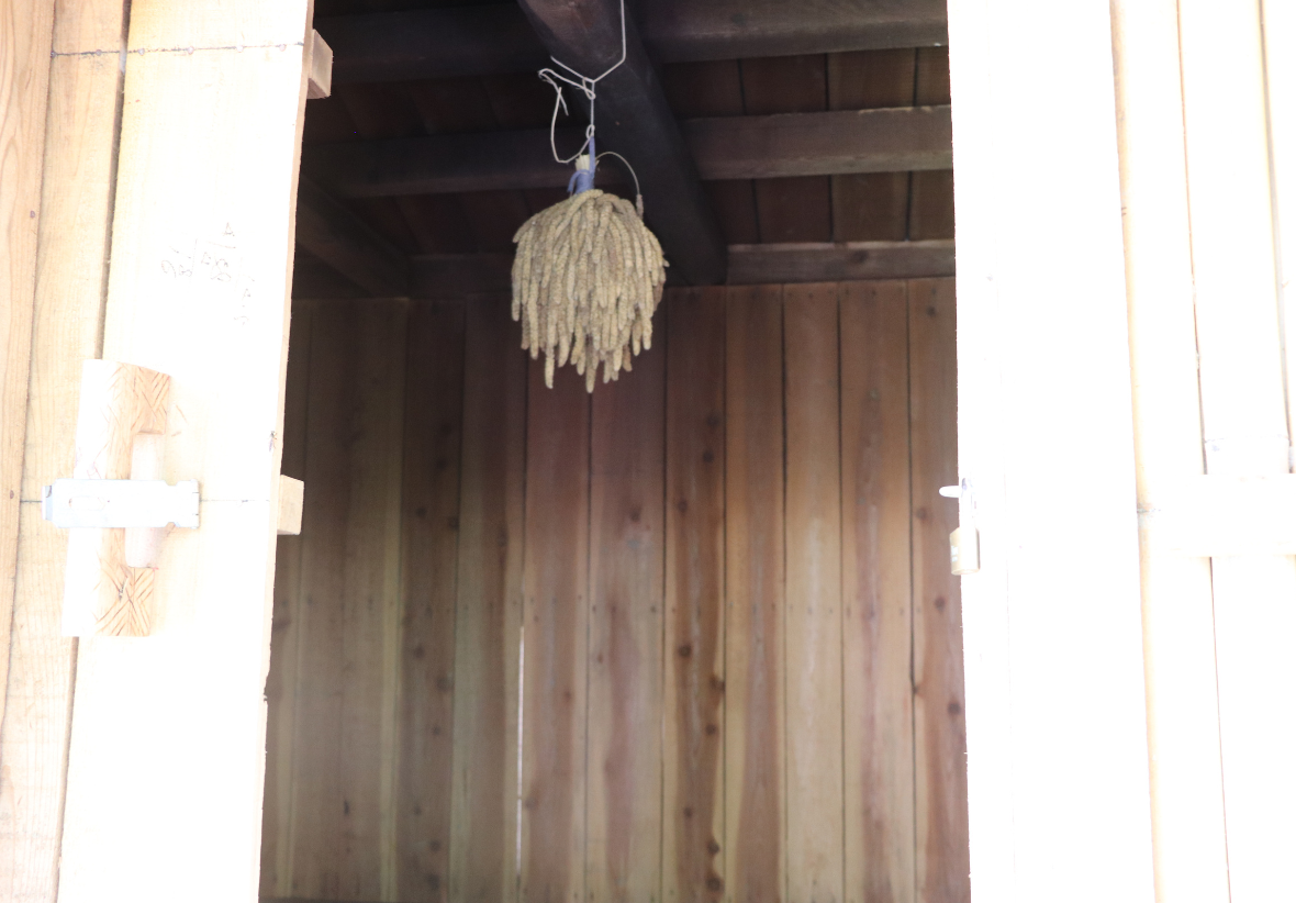 木造建築內部，白色木門敞開，門框左側裝飾有稻草十字架。天花板上懸掛著用繩子綁束的乾草裝飾品，營造出樸素的鄉村風格氛圍。