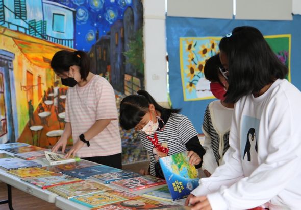 在美術教室內，一位成人教師和數名兒童圍著展示桌，一起欣賞和討論各式彩色繪本及藝術作品。牆面上裝飾著梵谷星月夜等藝術作品複製畫，營造充滿創意的學習環境。