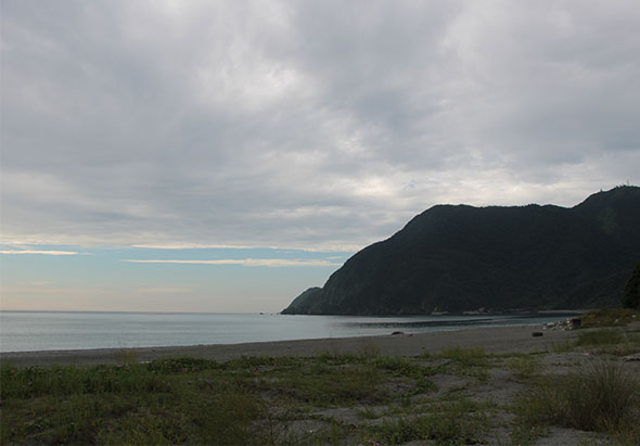 東澳海灘景觀，前景為沙灘和草叢，中景為寧靜的海面，遠景為陡峭的黑色山崖，天空布滿白雲，光線柔和，展現宜蘭沿海的自然風貌。