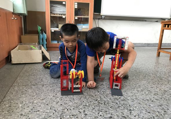 兩名穿著藍色制服的小學生在教室地板上，合作組裝紅色樂高建築模型。他們戴著獎牌，專注地進行STEAM教育活動。背景可見木製櫃子和教室設施。