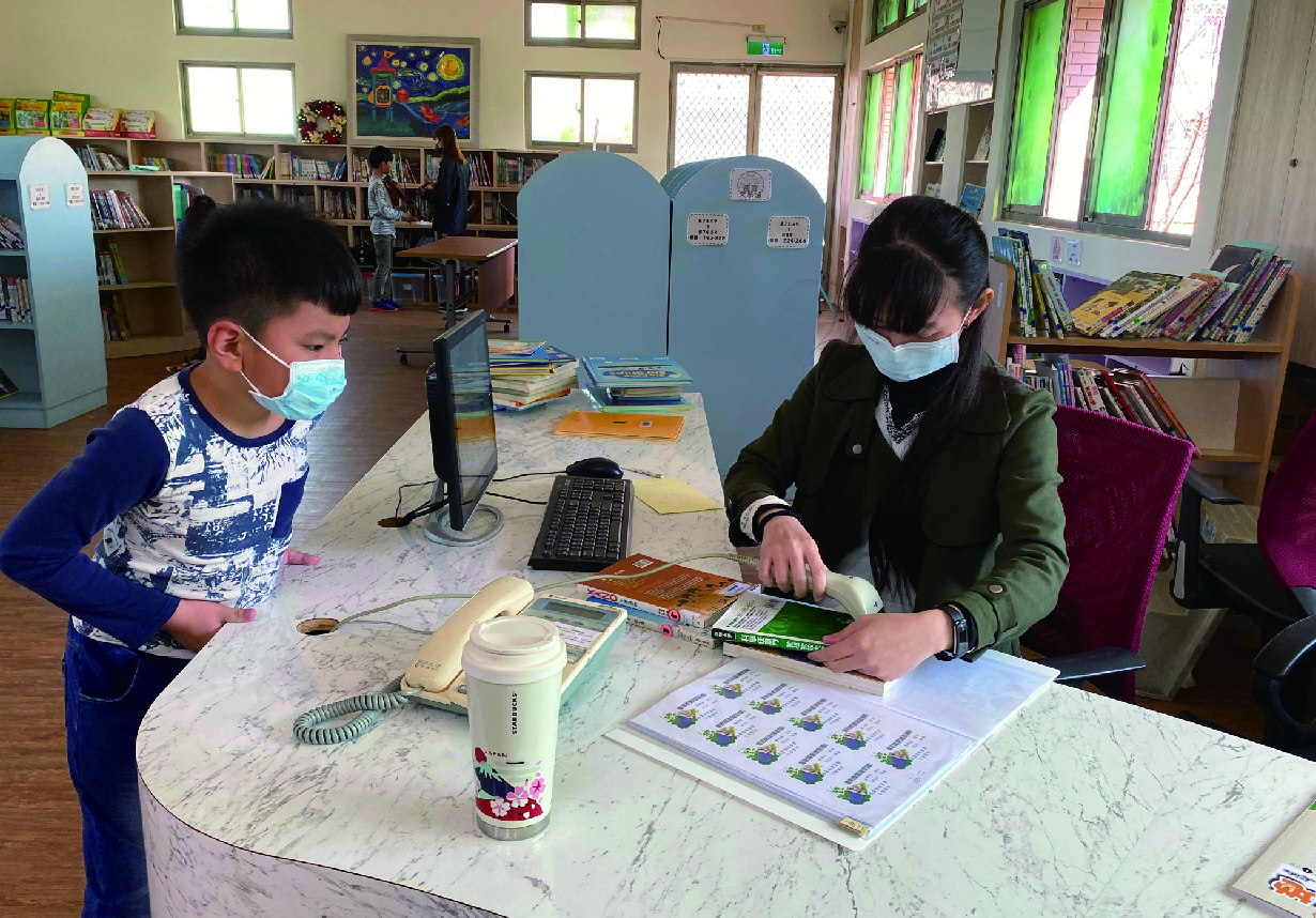 圖書館內兩名戴口罩的學童在圓形桌子前進行閱讀學習活動。左側男童穿著藍色上衣，右側女童穿著綠色外套，正查看書籍和學習材料。桌上放著飲料杯、書籍和列印文件。背景可見書架、其他學童和綠色門窗。