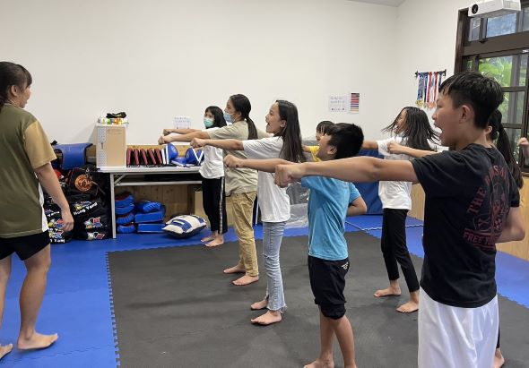 一群兒童在藍色運動墊上進行武術訓練，眾人站成一圈做出相同的防禦姿勢，伸展雙臂。教室內有儲物櫃和教學用具，窗戶外可見綠色植物。