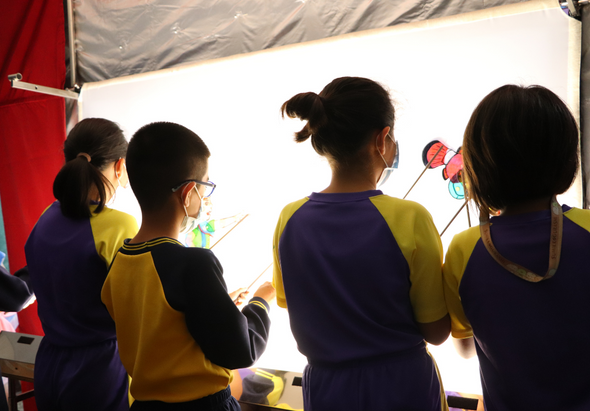 四名穿著藍黃相間運動服的學生在帳篷內，面對著發光的白色背景板，正在進行美勞或科學實驗活動。其中一人拿著畫筆或工具，專注於眼前的創作或觀察工作。