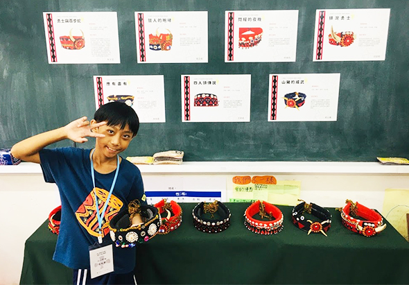 一位年輕學生在教室前展示傳統民族手工藝品，黑板上貼著七張中文標籤卡片，展示桌上擺放著多個紅黑色的傳統裝飾品和手鐲，呈現文化教育展示活動。