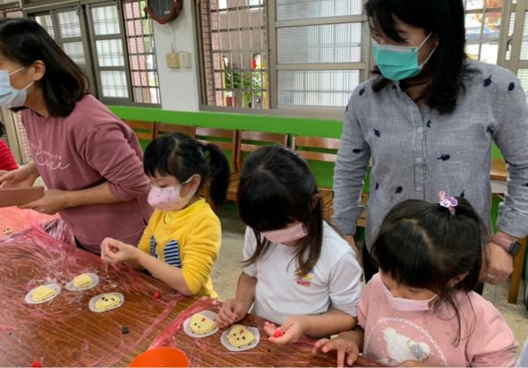 幼稚園教室內，兩位戴口罩的老師指導四位穿著粉色和黃色衣服的幼童，在木製桌上進行美勞活動，製作彩色裝飾盤。背景可見綠色黑板和玻璃窗。