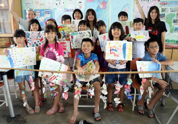 教室裡一群小學生展示自己的美術作品。學生們穿著整齊，有的拿著彩色畫作，有的展示手工藝品。前排學生坐著，後排站著，背景是布置滿教室佈告欄的牆面，呈現充滿創意的學習環境。
