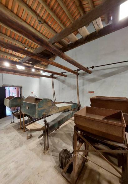 木製建築內部展示古代棺木和殮具，天花板為傳統木樑結構，地面為淺色木質地板。牆面白色，展示照明燈光充足。右側可見棕色木製棺木，左側為綠色古棺，中央懸掛傳統喪葬用具。此為新芳春古蹟室內陳設。