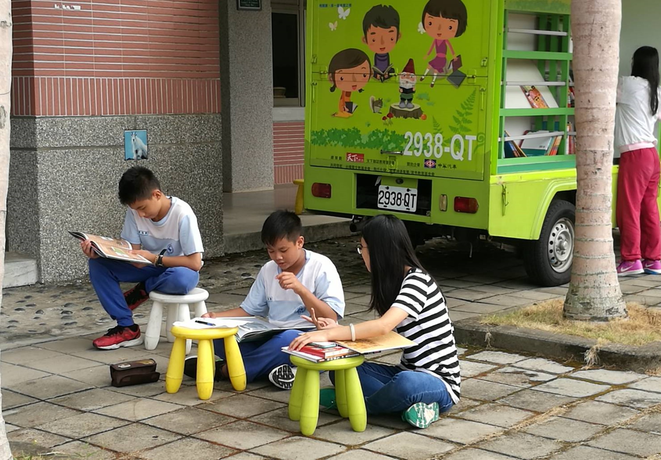 一位女性志工和三名小學生坐在戶外空地上閱讀。他們周圍放著黃綠色小凳子和書籍。背景是亮綠色的流動圖書館車，車身繪有卡通兒童圖案。這是一場推廣兒童閱讀的社區活動。