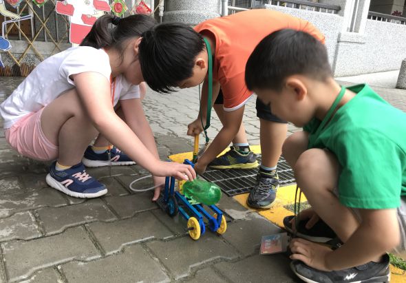 四位兒童蹲在戶外地面上，共同組裝或檢查一輛藍黃色的兒童玩具車。他們穿著不同顏色的衣服，認真地觀看和操作手中的玩具車零件，背景是建築物和停車場。
