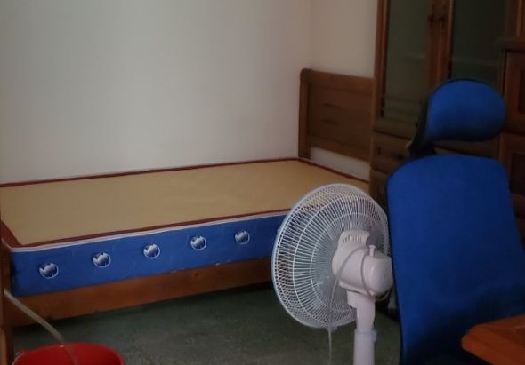 簡樸宿舍房間內，靠牆放置一張藍色邊框木製單人床，床頭有圓形通風口裝飾。房間角落擺放白色電風扇，右側放著藍色靠背椅子，牆面為白色，地板為綠色。