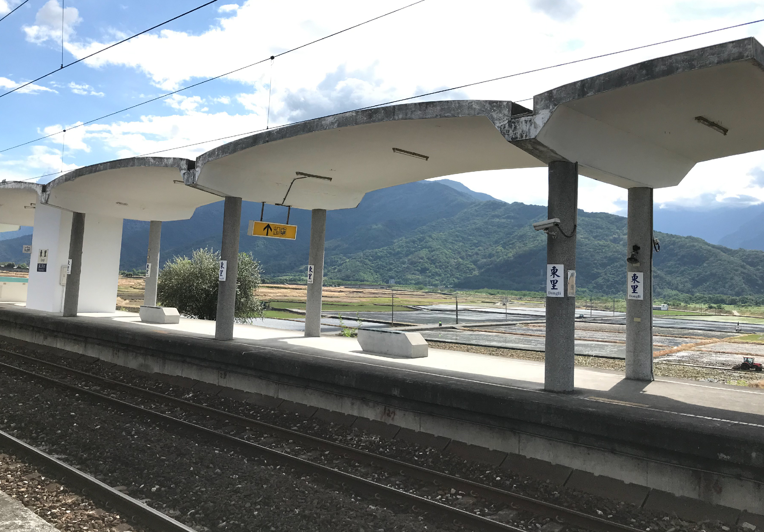 日本鄉村火車站月台，月台頂棚為白色混凝土結構，遠方可見綠色山脈和農田風景，月台邊有鐵軌，標示牌為日文。晴朗天氣，藍天白雲。