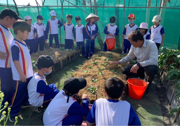 一位講師在溫室內向穿著校服的小學生教授園藝知識。學生們圍成一圈蹲坐著，認真觀看講師示範種植技巧。溫室內可見植物和紅色澆水桶，展現校園戶外教育活動的情景。