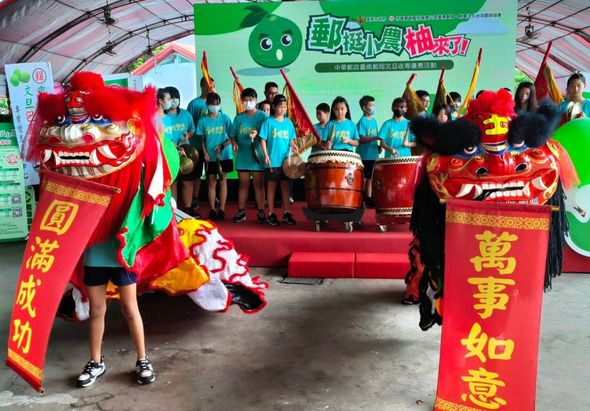 戶外活動現場，舞獅隊伍表演傳統舞獅，紅色獅頭兩側掛著金色祝福字幅。舞台上穿著藍色制服的參與者揮手歡呼，背景為綠色帳篷與活動標語，營造熱鬧慶典氛圍。