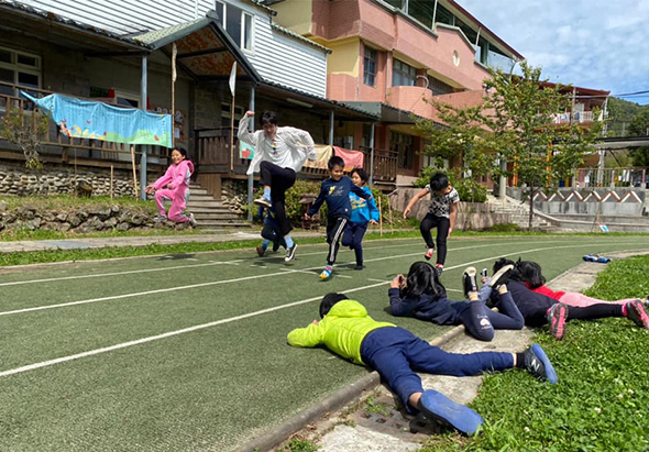 一群小學生在戶外運動場進行體育課，有學生在做伸展運動，有學生在進行跑步或移動訓練。背景是鄉村建築，綠色人工草皮運動場，晴朗天氣。