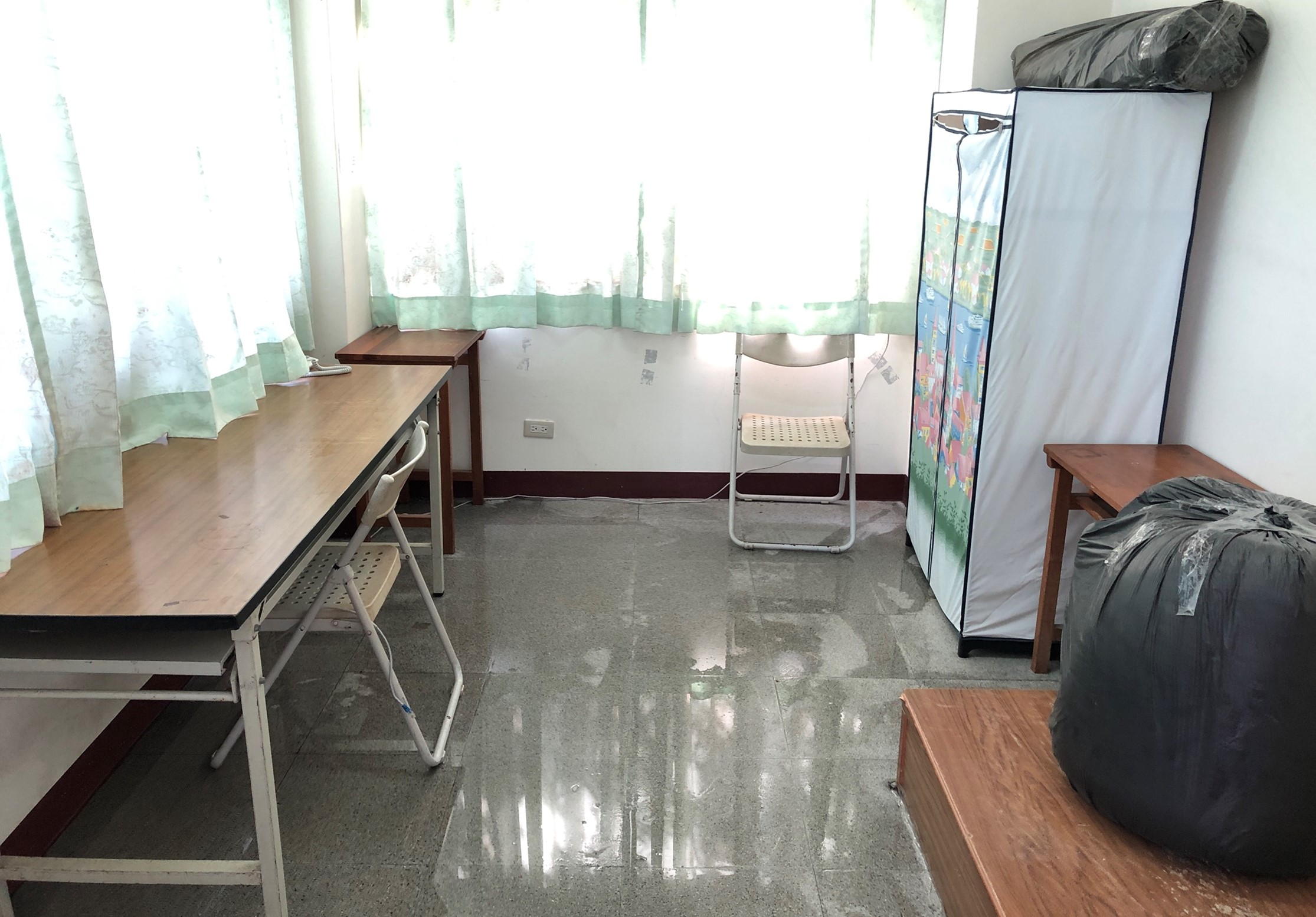 簡樸宿舍房間內景，地板濕潤反光，擺放有木製桌椅、折疊椅、衣架和打包行李。窗戶掛著綠色半透明窗簾，採光充足。牆面貼有電源開關，整體環境狹窄樸實。
