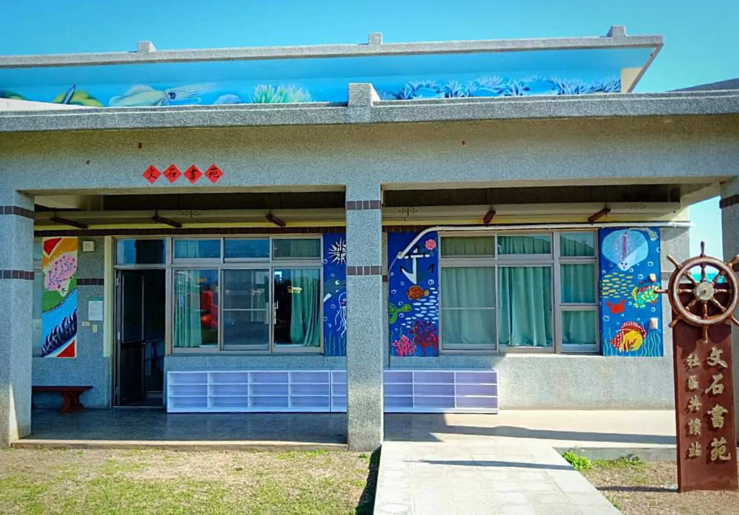 海邊主題的建築物正面，左右兩間店鋪裝飾著海洋生物壁畫，包括魚類、珊瑚和船舵等圖案。建築採用藍白色調，屋頂邊緣有海底場景浮雕。右側入口旁立有木製船舵裝飾和中文招牌。清晨陽光照亮整個建物。