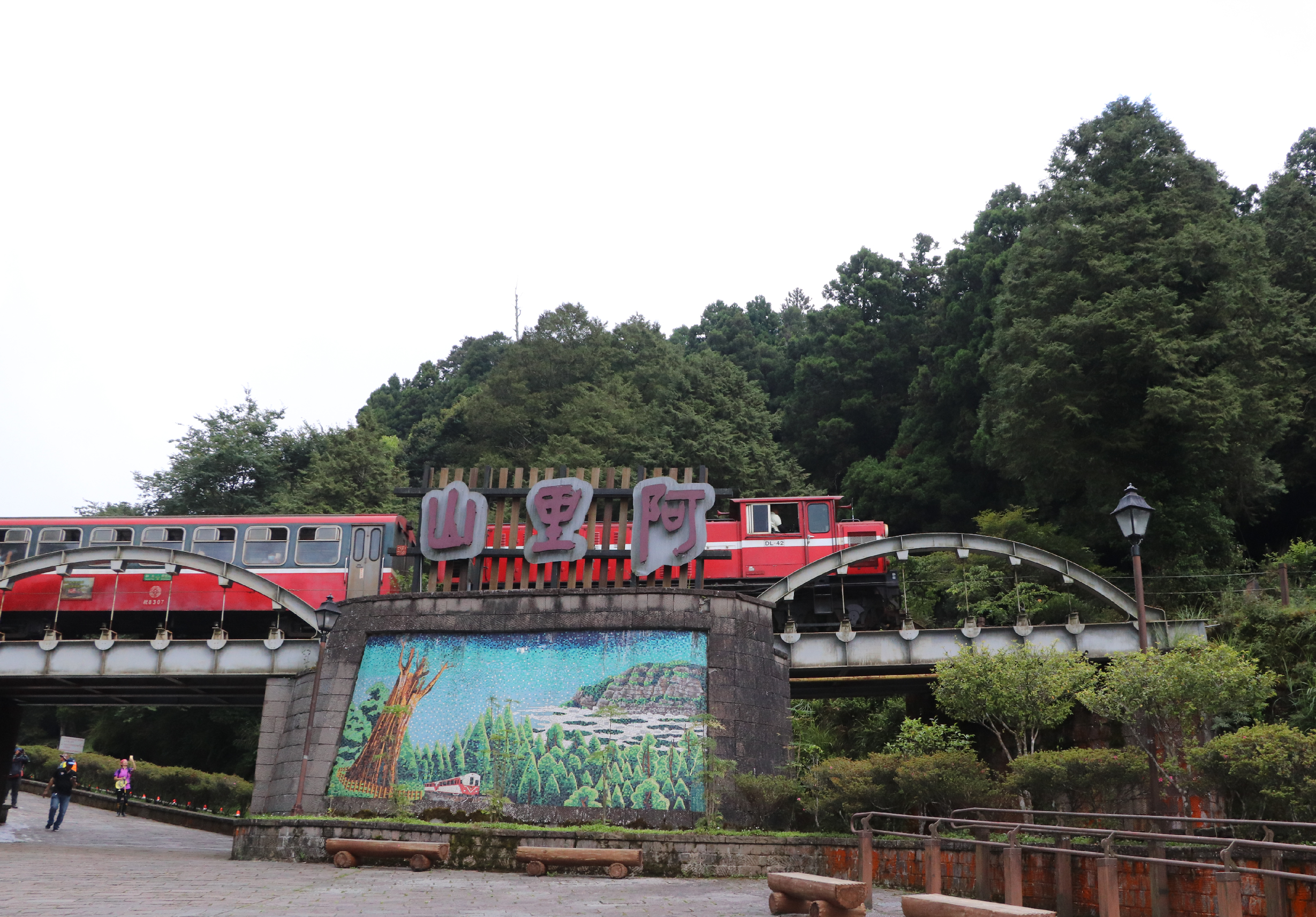 山寨風景區入口，紅色列車造型建築跨越石橋，橋墩上有紅色大字招牌，前方豎立著彩繪壁畫展示山林景致，遠處蒼鬱樹木，數位遊客漫步廣場。