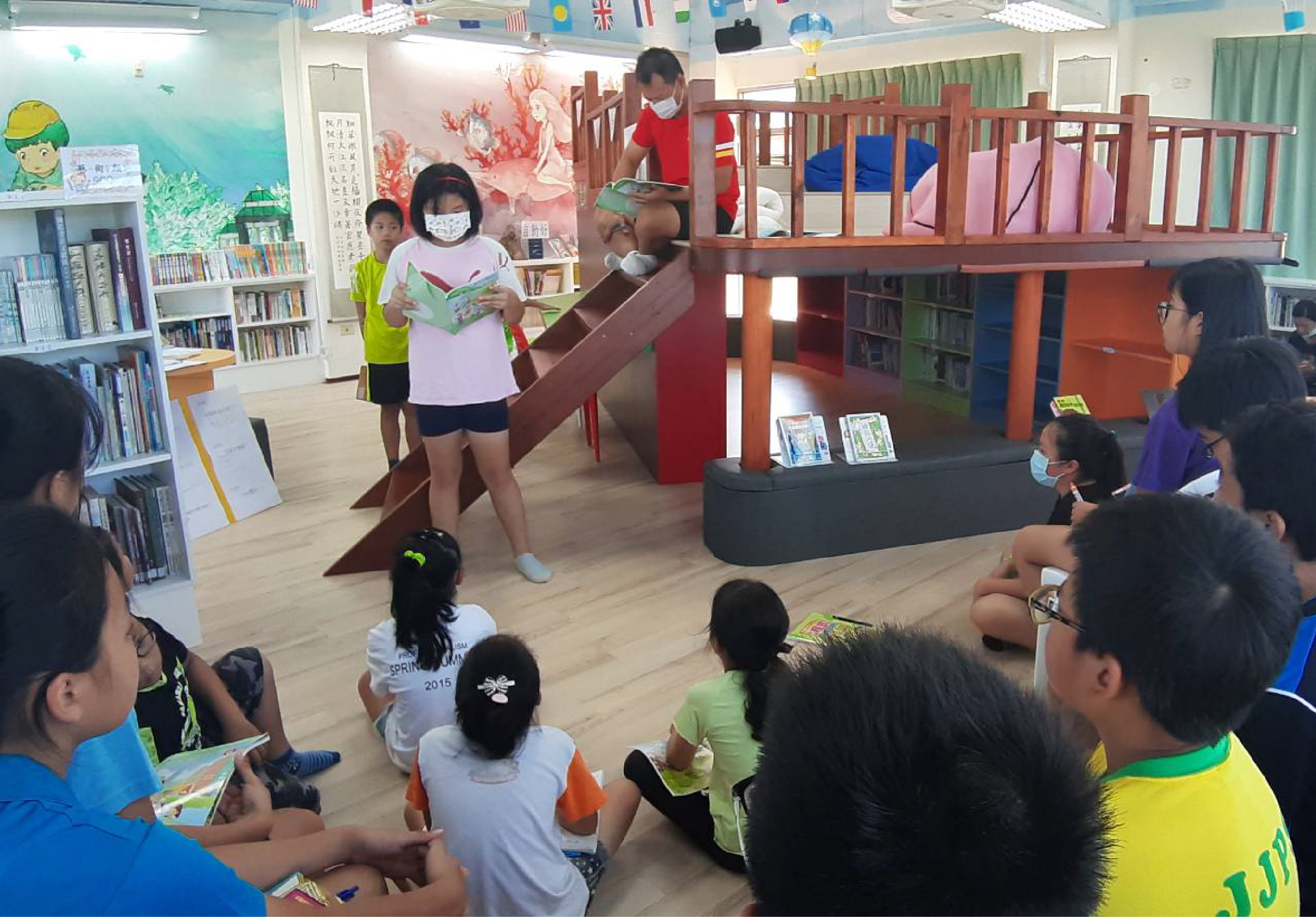 學校圖書館內，一位戴口罩的女學生站在木製閣樓前朗讀繪本，周圍圍坐著約十位穿著彩色運動服的小學生認真聆聽。背景為書架、彩色牆面及國旗裝飾。