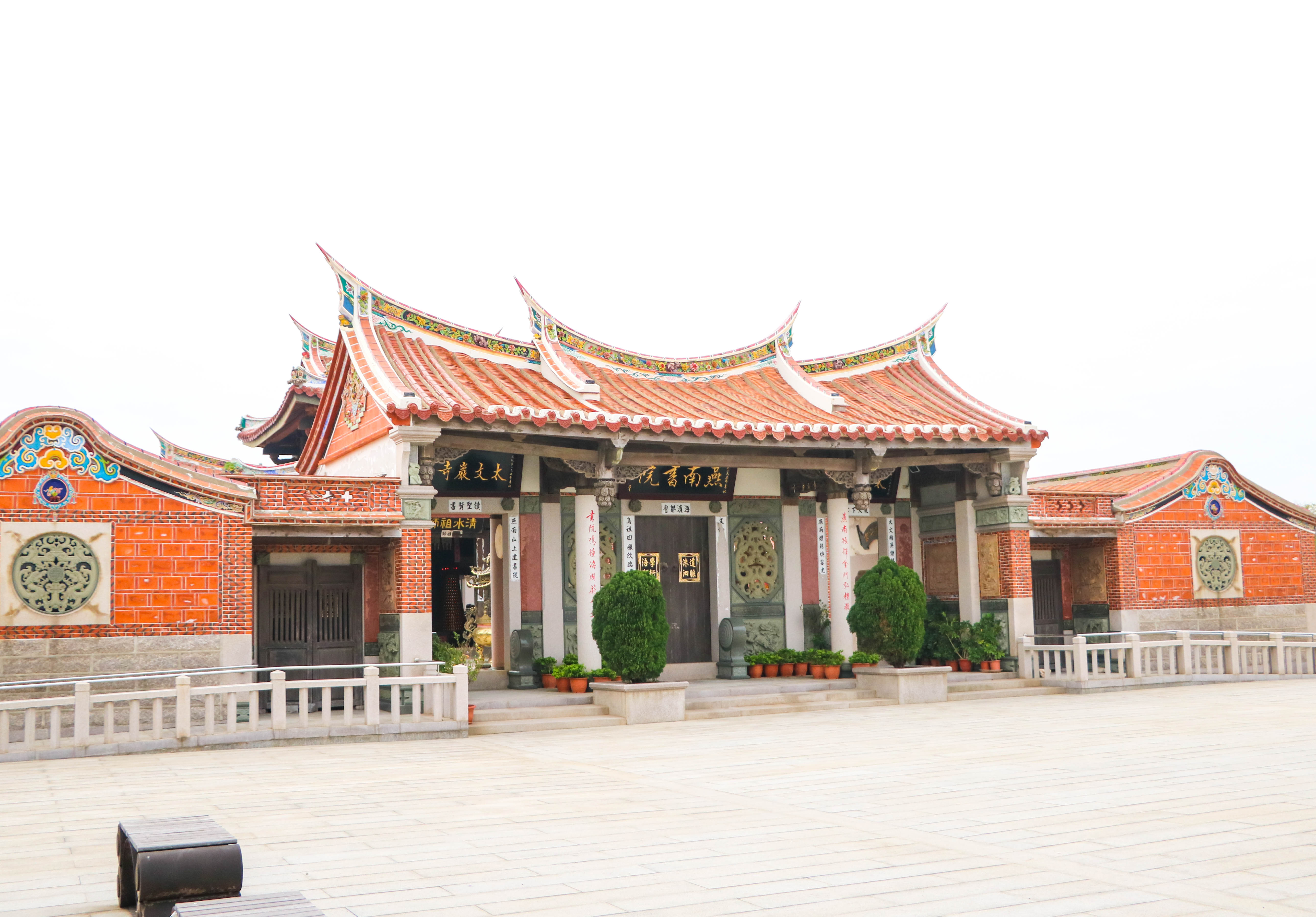 傳統中式廟宇建築，紅磚牆面配紅瓦屋頂，屋脊上翹起，裝飾著金色雕紋。廟門兩側有柱子和對聯，前方擺放陶製花盆和綠色植物。廣場鋪設淺色石板，周圍有白色欄杆。建築典雅莊嚴，展現東方傳統建築特色。