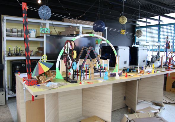 工作坊內展示一座大型木製模型建築，配有綠色拱門、彩色風車和懸掛球體裝飾。木製工作台上散放工具和材料，背景有白色櫃子、螢幕和工業風格天花板，呈現創意製作空間。