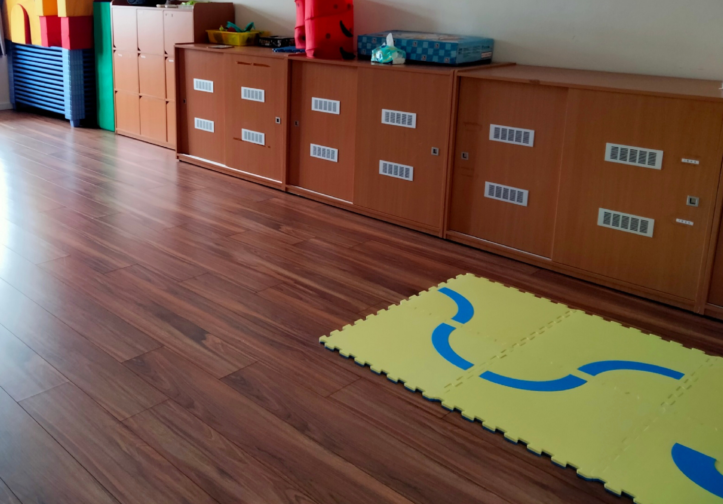 幼兒園教室內，木製地板上鋪著黃色泡沫墊，上面有藍色波浪圖案。背景為棕色儲物櫃和彩色教具，窗明几淨的學習空間。