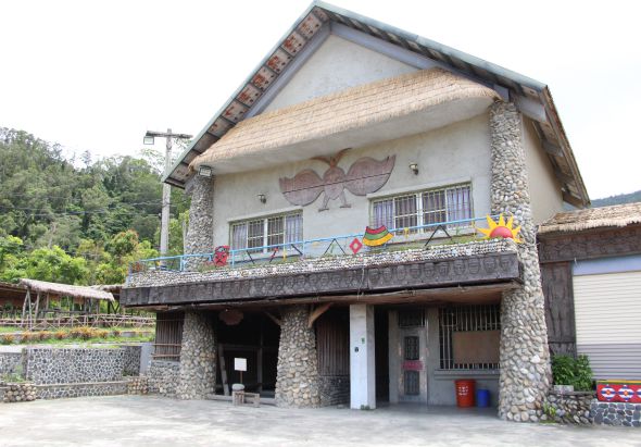 石造鄉村建築，二層樓房設有寬敞陽台，裝飾著紅花和黃色裝飾品。一樓為開放式商店或服務區，周圍綠樹環繞，右側停放白色車輛，背景可見電線桿。