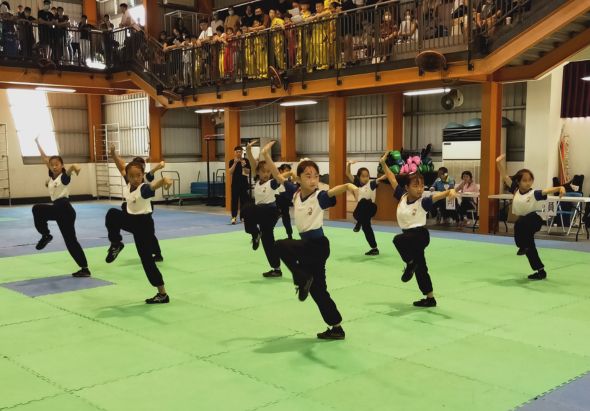 室內體操館中，穿著黑白運動服的學生們在綠色墊子上整齊排列，做出同步的跳躍動作。二樓觀眾席坐滿了觀看者，木製建築結構清晰可見，展現了一場集體運動表演或訓練課程。