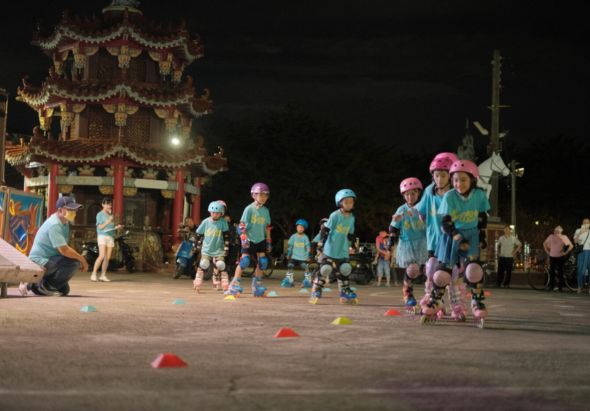 夜間在傳統中式廟宇廣場，眾多身穿藍色和粉紅色衣服、戴著頭盔的兒童進行直排輪訓練。場地上擺放著紅色和黃色的訓練路標。成人教練在一旁指導。背景為金碧輝煌的兩層飛檐廟宇和紀念碑。