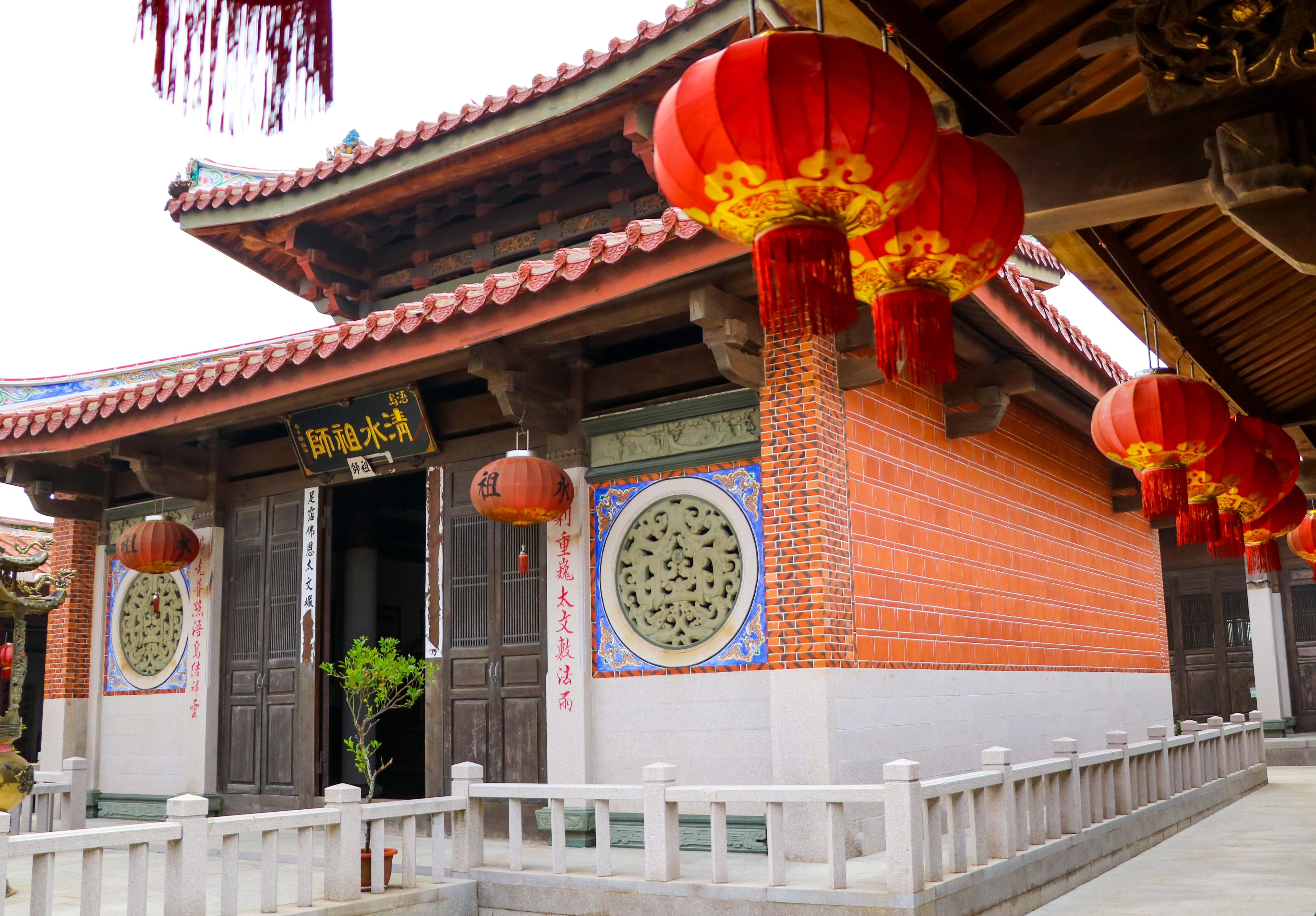 傳統中式廟宇建築，紅瓦屋頂與紅磚牆面，懸掛大紅燈籠，門前裝飾圓形玉石雕刻，白色石欄杆環繞，融合中華建築特色與文化元素。