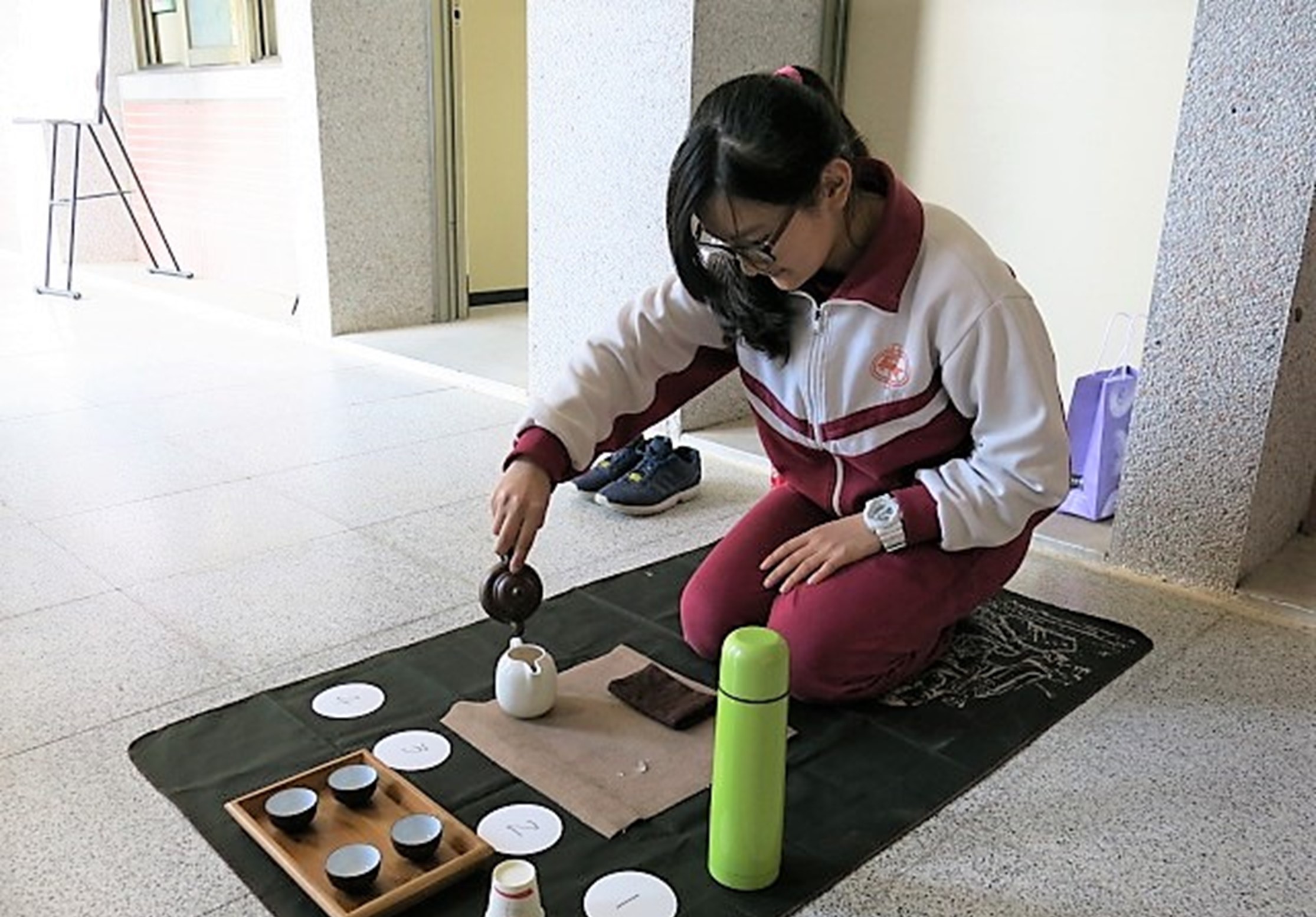 穿著校服的女學生在室內地墊上進行茶藝表演，她正在倒茶。地墊上擺放著茶具、茶杯、綠色保溫瓶和其他茶道用具，展示傳統茶文化活動。