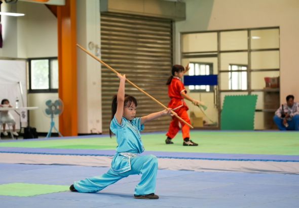 室內體操館中，穿著藍色運動服的小女孩在墊子上做武術動作，揮舞著繩子。背景有穿紅色服裝的另一名兒童和其他學員在進行訓練活動。