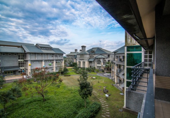 從高樓層陽台俯瞰現代住宅社區，綠色草坪中央有步道和植栽，周圍環繞多棟白色及灰色建築，藍天白雲映襯寧靜的居住環境。