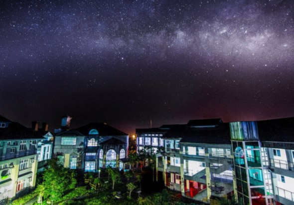 夜晚城市街景，多棟建築物亮著窗燈，上方是壯觀的銀河星空，星辰密佈，展現城市與自然的對比，充滿寧靜的夜間氛圍。