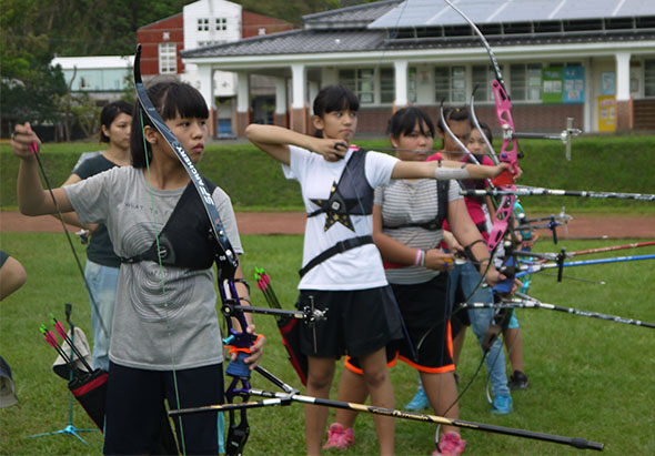 四名學生在校園草地上進行射箭活動，她們手持五顏六色的弓箭，擺出準備射擊的姿勢。背景是校舍建築和綠色環境，展現戶外運動教學情景。