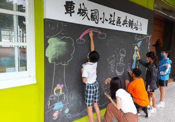 一群學童在黃綠色牆面的黑板上進行創意塗鴉活動。黑板上方有中文書法標題，學童們用粉筆畫出樹木、人物、動物等圖案。穿著白襯衫和格子褲的學童站在板前揮灑創意，其他學童則在旁邊觀看和參與。這是一場互動式的校園藝術教育活動。