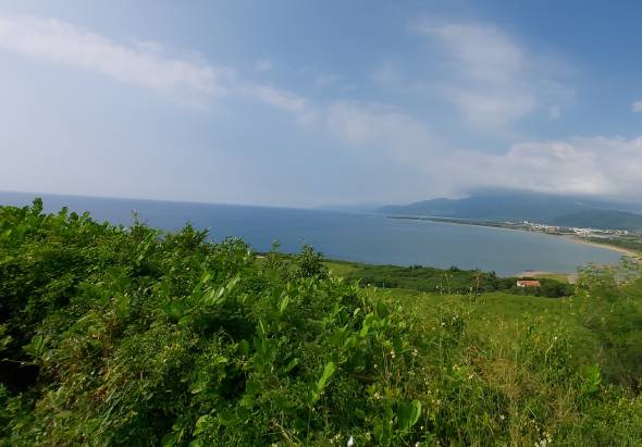 從山坡上俯瞰遼闊的海灣景觀，青綠色植被覆蓋前景，遠處藍色海面與對岸山脈相接，沿岸可見建築物和沙灘，天空晴朗飄有白雲。